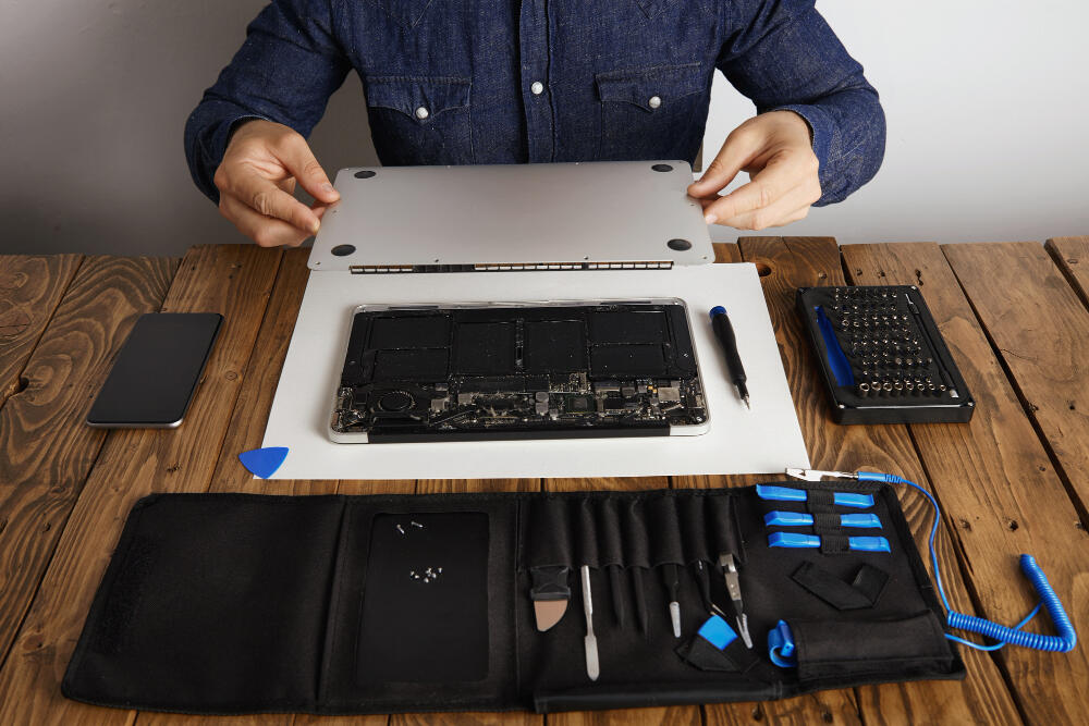 Mãos de técnico utilizando uma espátula plástica azul para abrir a tampa traseira de um notebook prata sobre uma bancada de madeira.